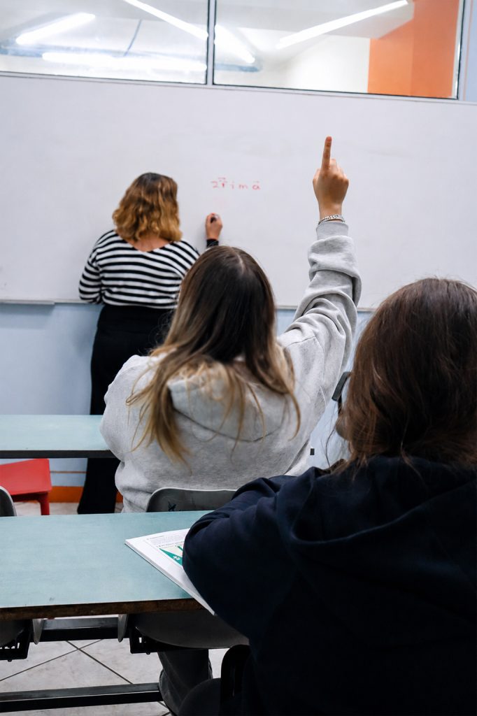 Μαθήτρια σηκώνει το χέρι μέσα σε φωτεινή αίθουσα διδασκαλίας, ενώ η καθηγήτρια γράφει στον πίνακα.